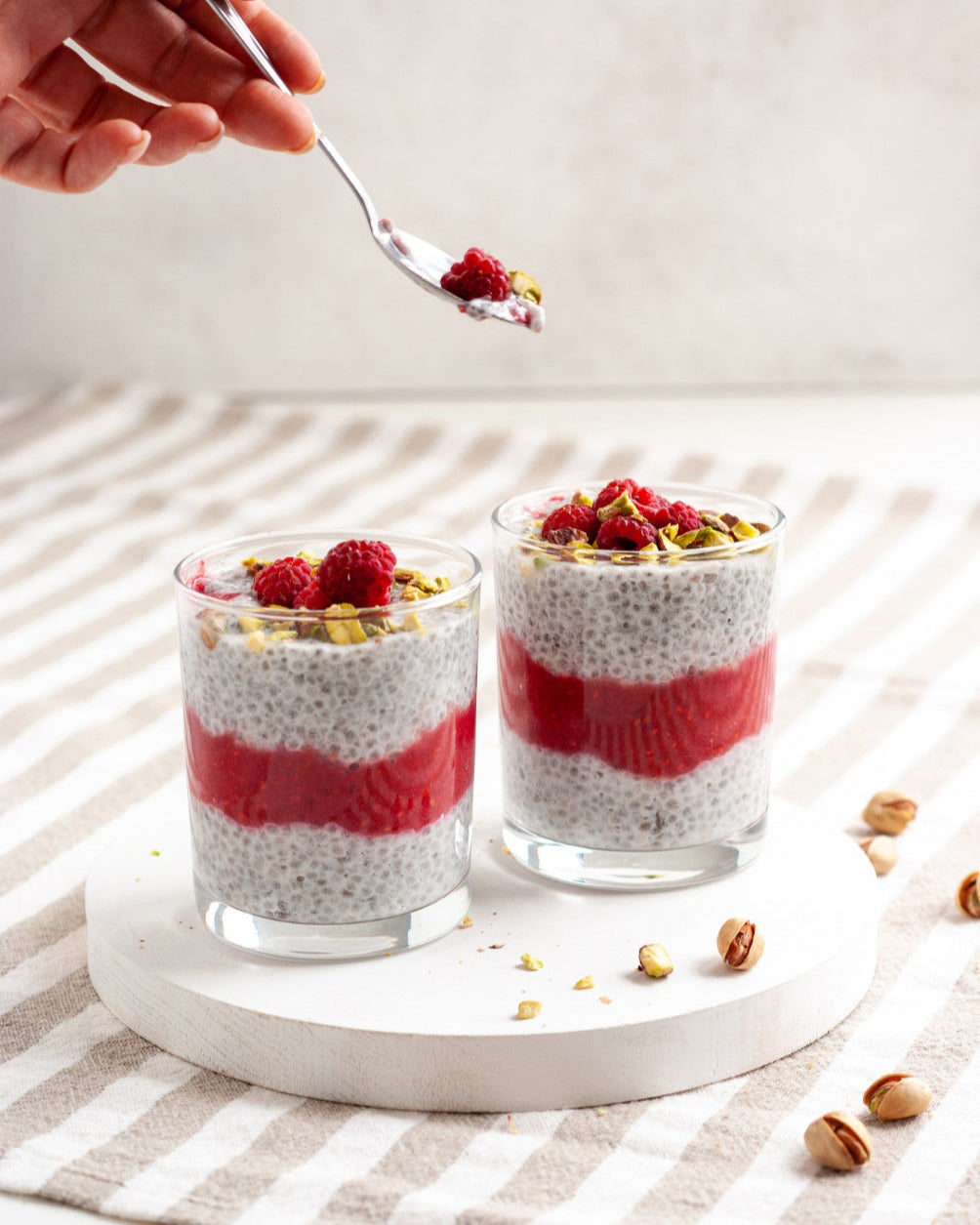 Two glasses of chia pudding with raspberries and pistachios on a striped cloth.