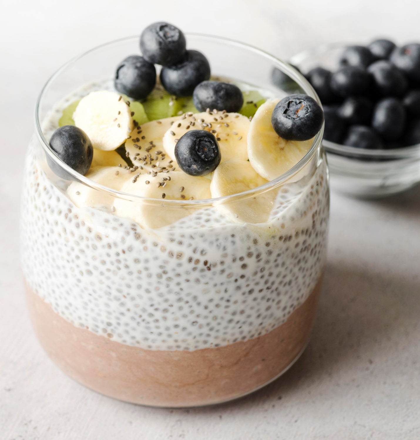 Glass jar with chia pudding topped with bananas, blueberries, and kiwi on a light surface.